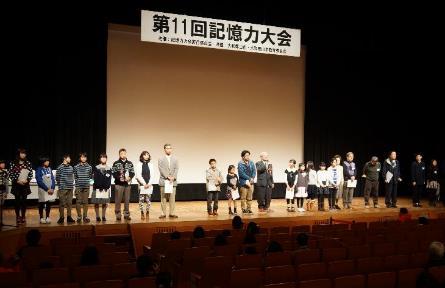 「第11回記憶力大会」と書かれた横断幕と白いスクリーンのある舞台上に並び立つ白い紙を持った大勢の男女の大人や子供たちの写真