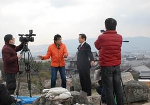 石垣の上に立つオレンジのジャンパーを着た男性タレントとスーツ姿の男性をビデオカメラで撮影する2人の男性の写真
