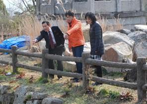 石垣の上の柵のそばに立つ遠くに向けて指をさすオレンジのジャンパーを着た男性とスーツ姿の男性と2人の男性の右隣で直立している黒いジャンパーを着た女性の写真