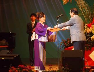 舞台上の黒いスタンドマイクのそばでグレーのジャケット姿の男性から白い紙を受け取る紫色の着物姿の女性の写真