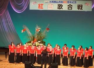 紅白歌合戦と書かれた横断幕のある舞台上に並び立つ赤い半袖の服に黒いスカートをはいた女性たちの写真