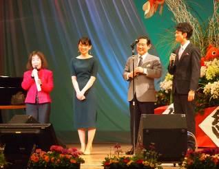黒いスタンドマイクのある舞台上に立つ2人のスーツ姿の男性と男性たちの左側に立つ青いワンピースの姿の女性とマイクを持つピンク色のスーツ姿の女性の写真