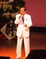 「絆」と書かれた台の上に花束のある舞台上で白いスーツを着て右手にマイクを持っている中年男性の写真