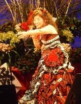 舞台上で赤色を基調としたドレスを着て右手にマイクを持ち左手で指をさす仕草をする中年女性の写真