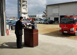 防災センター内の停車している赤い消防車の前で男性とスーツ姿の男性が白い紙を受け渡ししている様子の写真