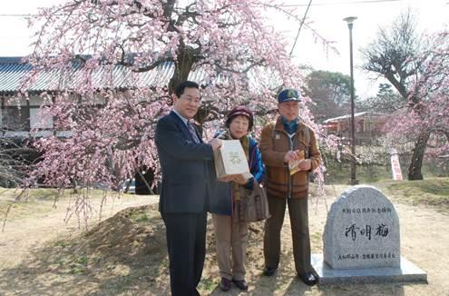 「清明梅」と書かれた石碑としだれ桜のそばに立ち「器」と書かれた紙袋を持つスーツ姿の男性と高齢男女2人の集合写真