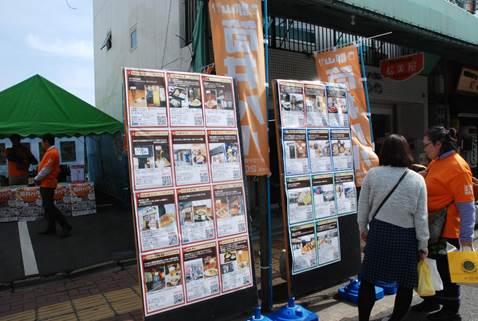 日本の筒井バルと書かれたオレンジ色ののぼりのそばにある郡山筒井バルに参加する11の店舗の紹介写真が貼られた看板を見つめる二人の女性の写真