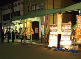 道路わきに立ち並ぶ建物のそばで明かりが照らされた筒井バルに参加する店舗一覧の2つの看板と筒井バルと書かれたオレンジ色ののぼりが立ち並ぶ夜の写真