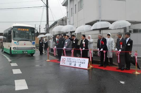 コミュニティバス開通式と書かれた看板のそばで各々後ろに立つ人物に傘をさしてもらっている設置された紅白のテープの前に立つスーツ姿の男性たちと男性たちの近くにある白と緑色のツートンカラーのバスの写真