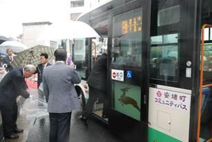 ドアが開いたコミュニティバスに乗り込むスーツ姿の男性とそばに立つビニール傘を持った男性たちの写真