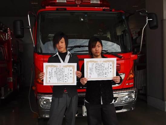 赤い消防車の前に並び立ち両手に持った感謝状を掲げる2人の少年の写真