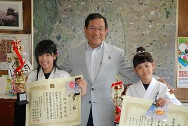 大きな地図が壁に貼られている室内に立つグレーのスーツを着た市長と市長の両隣に立ち賞状とトロフィーを手にする空手の胴着を着た2人の女の子の写真