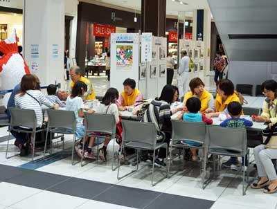 イオンモール大和郡山店内にある白い長机に向き合うように座る黄色いベストを羽織った男女4人と女性と男女の子どもたちの写真
