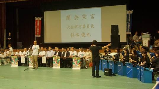 「開会宣言大会実行委員長杉本俊也」の文字が投影された大きなスクリーンを背に着席する男女の人々の前でスタンドマイクの前に立つTシャツを着た男性と右脇の青い机に座る男女の黒い服を着た人々の写真
