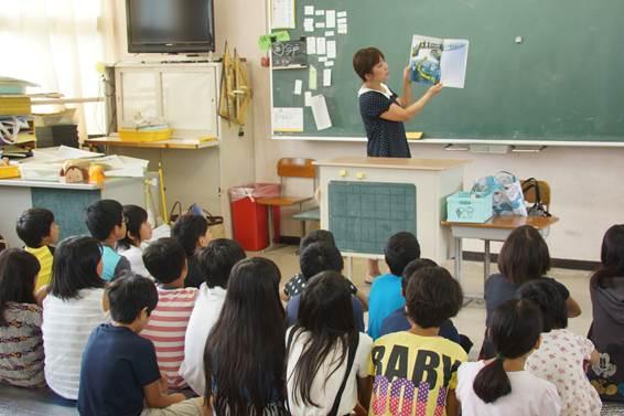 左脇にテレビがある白い教室で集まり座る男女の子どもたちを前に黒板を背に教壇のそばに立ち両手で絵本を持つ女性の写真