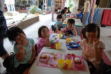 幼稚園の外のカレーライスの入った弁当箱が並んだテーブルに座る笑顔の男女の園児たちの写真