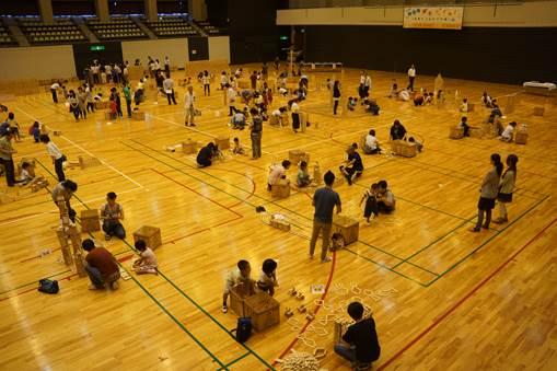 床に散乱する木製のブロック板と点在するブロック板の入った箱に集まる人々がいる体育館の室内の写真
