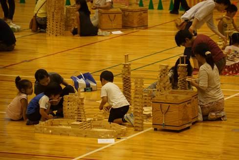 体育館の床の上で積み重なった木製のブロック板とブロック板を積み上げている男女の子どもたちと女性の写真