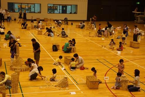 体育館の床の上でグループ分けされた男女の子どもたちと大人たちが木製のブロック板を積み重ねている写真