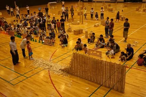 体育館の床の上で半分位崩れた積み重なる巨大な木製のブロック板のそばに立つ子どもと男女の大人と遠くから様子を見つめる大勢の参加者たちの写真