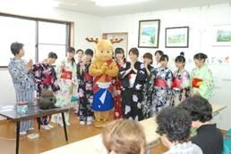 室内で長机に着席する来場者たちを前に両手裏を重ね合わせている奈良県のゆるキャラ「せんとくん」と大勢の着物姿の女性が集まる集合写真