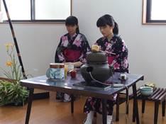 室内の窓辺で黒い茶釜や陶器が置かれた黒いテーブルに着席している黒地にピンク色の花模様の着物を着た2人の女性の写真