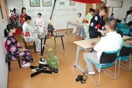 赤い大きな和傘が飾られた部屋で着席する着物姿の女性たちや長机に座っている来場者の男性にお茶をふるまう2人の着物姿の女性の写真