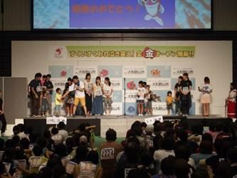 全国金魚すくい選手権大会の奈良県の予選会場内のステージに集まる男女の子どもや大人たちと着席して彼らを見つめる大勢の人々の写真