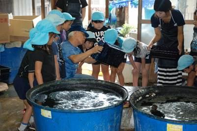 水が入った二つの大きな桶の傍で道具を持って説明する西井さんに集まる園児たちの写真