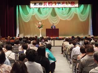大きな花が飾られた舞台上で話すスーツを着た男性と椅子に座っている多くの来場者の後ろ姿の写真