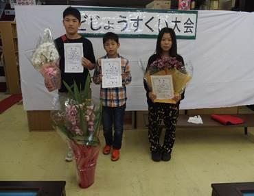どじょうすくい大会と書かれた横断幕のある白い壁を背景に賞状と花束を持って立つ女の子と男の子と2人の真ん中で両手に賞状をもつ男の子の写真