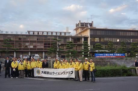 雲が浮かぶ空の下市役所の建物を背景に「大和郡山ライオンズクラブ」と書かれた横断幕を持つ黄色いジャンパーやスーツを着た男女たちの集合写真