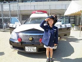 屋外に止まっている両側のドアが開いた奈良県警のパトカーの前で女性警察官の制服を着て右手で敬礼をする女の子の写真
