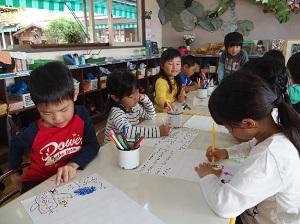 大きな窓と茶色い棚のある室内にある白いテーブルの上で絵や文字を書く男女の子どもたちの写真