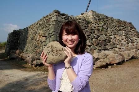 青空の下石垣を背景に文字が書き込まれた石を両手で持ち笑みを浮かべる女性の写真