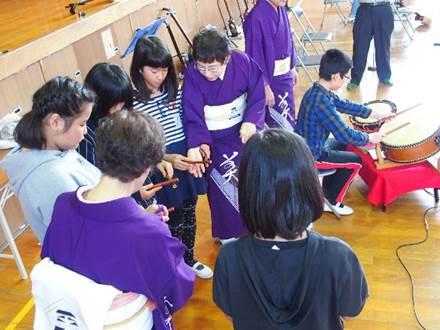 木の床の室内にいる紫色の着物を着た3人の女性と4人の女の子と両手にバチを持ち2つの太鼓の前に座る男の子の写真