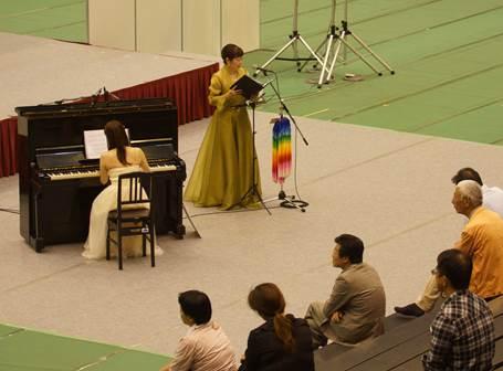 室内の緑色の床に敷かれた白い布の上でピアノを弾く白いドレス姿の女性と千羽鶴で飾ったスタンドマイクの前に黒い楽譜を両手に持つ女性と席に座っている5人の男女の写真