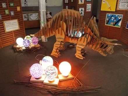 室内の中心にダンボールの板を組み合わせて作られた大きなトリケラトプスのオブジェと2か所に集められている4つの色とりどりの光る丸い形をしたランプの写真