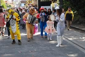 道路を虎などの動物の仮装をしてアコーディオンなどの楽器を演奏しながら練り歩く集団と集団のそばを行きかう人々の写真