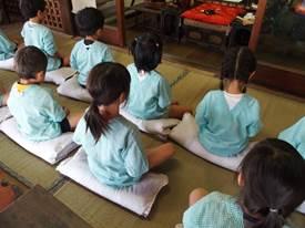 お寺の室内の畳の上で水色のスモックを着た園児たちが座布団の上で座禅を組んでいる写真