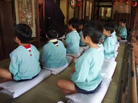 お寺の室内の畳の上で水色のスモックを着た園児たちが座布団の上で座禅を組んでいる様子を左から見た写真
