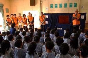 青い布に覆われた閉じられた紙芝居の本がある幼稚園の白い室内で水色のスモックを着た園児たちの前に立つオレンジ色のベストを着た男女たちの写真