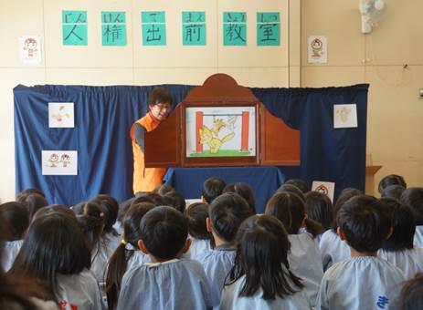 幼稚園の白い室内で水色のスモックを着た園児たちの前に立ち開いた紙芝居の絵本を見せるオレンジ色のベストを着た女性の写真