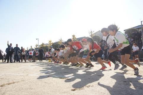 青空のもと運動場で色とりどりのたすきを掛けた子どもたちが横一列に並び走り出そうと体を前のめりにしている写真