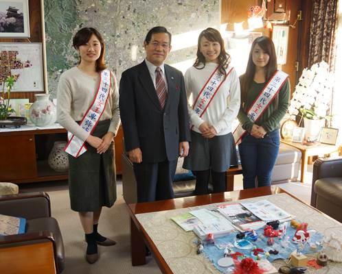壁に大きな地図のある市長室のテーブルの前に立つ紅白のたすきを掛けた3人の女性とスーツ姿の市長の集合写真