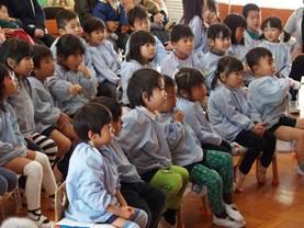 幼稚園の室内で集まるように椅子に座っている水色のスモックを着た男女の園児たちの写真
