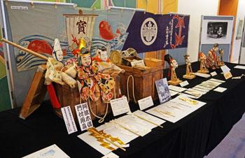 黒い布が敷かれたテーブルに宝船を模した七福神の人形が展示されている様子の写真