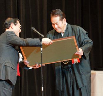 黒いスーツの男性から大きな額縁を手渡される黒い着物を着た受賞者の写真