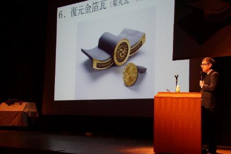 登壇した講師がスクリーンで、金箔が張られた瓦の説明を行っている処の写真