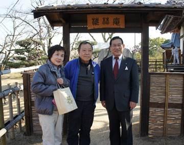 木と竹で作られた玄関の前で、スーツを着た男性から記念品を貰っている夫婦の写真